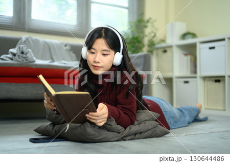 Young woman lying on the floor with headphones, reading a book and relaxing at home 130644486