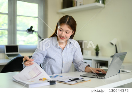 Businesswoman analyzing financial reports and typing on laptop in modern office Businesswoman analyzing financial reports and typing on laptop in modern office 130644573