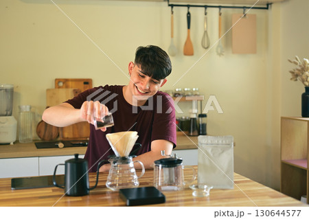 Smiling young man pouring ground coffee into a dripper for manual brewing at home Smiling young man pouring ground coffee into a dripper for manual brewing at home 130644577