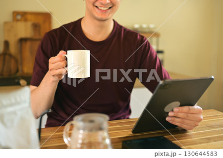 Smiling man holding a white coffee mug while using a digital tablet at home kitchen 130644583