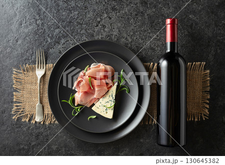 Bottle of red wine with blue cheese, prosciutto, and rosemary on a black table. 130645382