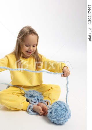 Pretty little girl in yellow clothes plays with ball of blue thread. Child model with long blond hair sits in studio on light background. High quality photo 130647467