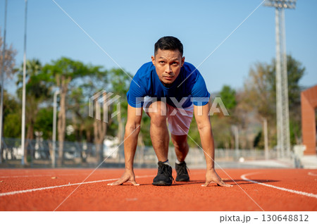 An asian man is crouching in a set position ready to run on the racetrack. Outdoor exercise. An asian man is crouching in a set position ready to run on the racetrack. Outdoor exercise. 130648812
