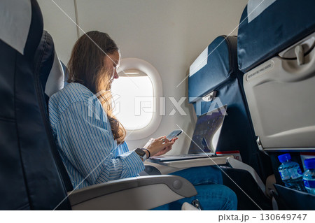 Young woman working on a laptop during a flight, seated by the airplane window. Remote work, travel productivity, and digital lifestyle on the go 130649747