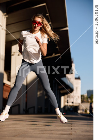 Athletic woman exercising outdoors under bright sunlight wearing sportswear and sunglasses 130650138