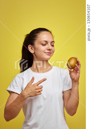 Happy woman in white t-shirt holding kiwi on studio yellow background 130654107
