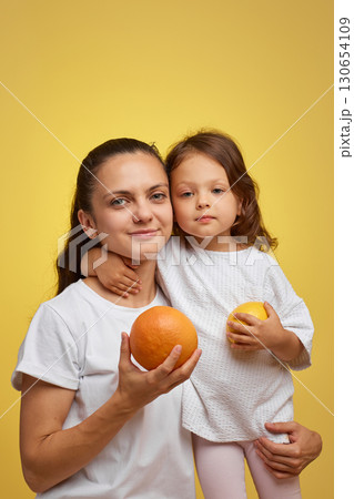 Cheerful little child girl with mother holding lemon and grapefruit on yellow background. happy family having fun together 130654109