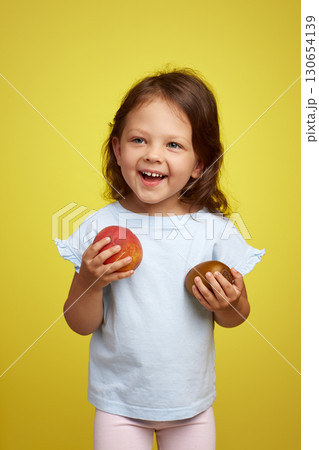 cute smiling Caucasian little child girl holding kiwi and peach on yellow background cute smiling Caucasian little child girl holding kiwi and peach on yellow background 130654139