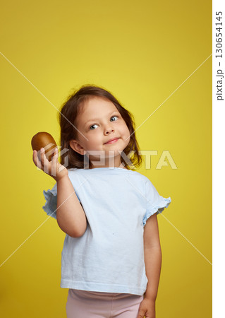 cute Caucasian little child girl holding kiwi fruit on yellow background 130654145