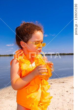 Girl in orange swimsuit drinking juice with yellow straw on beach with playful face Girl in orange swimsuit drinking juice with yellow straw on beach with playful face 130654190
