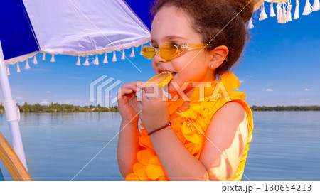Smiling girl biting lemon slice in yellow swimsuit under umbrella at beach 130654213