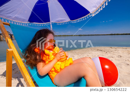 Girl in orange swimsuit sipping citrus drink under beach umbrella with beach ball 130654222