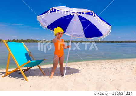 Kid in yellow swim cap with orange swimsuit holding umbrella on beach with confident face 130654224