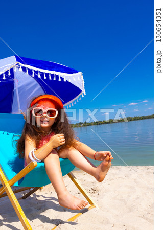 Girl in sunglasses sitting on deck chair at beach umbrella with joyful smile Girl in sunglasses sitting on deck chair at beach umbrella with joyful smile 130654351