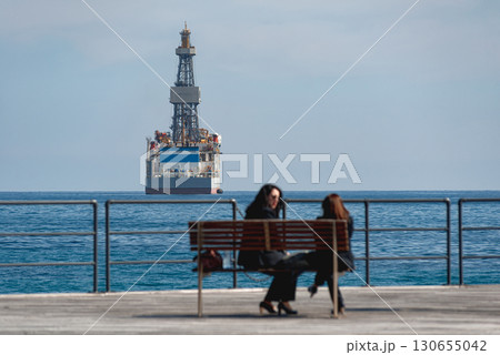 Two women sitting on bench with Drilling Vessel in background. Akrotiri bay, Limassol, Cyprus Two women sitting on bench with Drilling Vessel in background. Akrotiri bay, Limassol, Cyprus 130655042