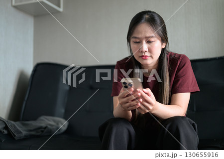 Young Thai woman sitting on sofa using smartphone at home, lifestyle portrait Young Thai woman sitting on sofa using smartphone at home, lifestyle portrait 130655916