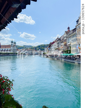 Breathtaking view of historic buildings lining a serene river under a clear blue sky. Bright flowers adorn the riverbank, while people enjoy outdoor dining by the water 130657624