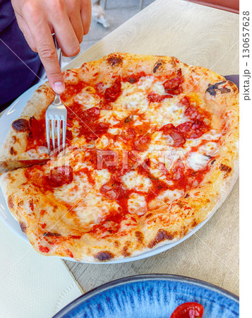 Pizza being cut with a fork and knife on a plate, displaying a crispy crust and melted cheese. The scene captures a moment of savoring a delicious meal. Perfect for food-themed visuals. 130657628