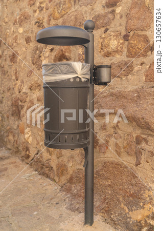 Urban trash can equipped with a plastic liner against a textured stone wall. The sturdy design and practical placement make it ideal for maintaining cleanliness in busy public spaces. Urban trash can equipped with a plastic liner against a textured stone wall. The sturdy design and practical placement make it ideal for maintaining cleanliness in busy public spaces. 130657634