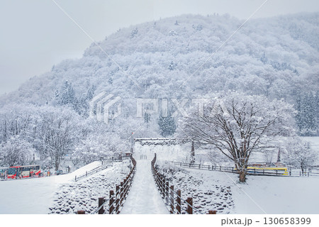 雪の白川郷・であい橋を渡る人々、庄川に架かる吊り橋と白銀の世界遺産集落 130658399