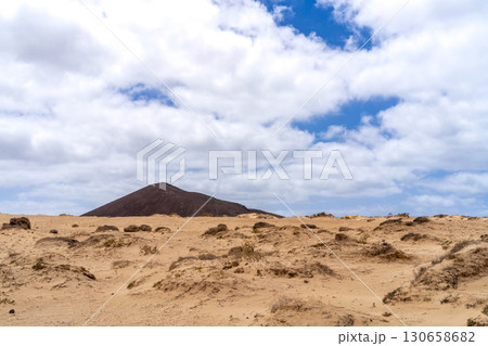 A scenic view of La Graciosa, showing a volcanic hill over sand dunes under a cloudy blue sky. 130658682