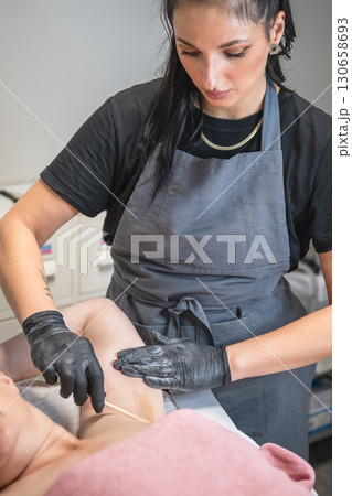 Beautician applying wax with wooden spatula on female armpit during professional hair removal procedure 130658693