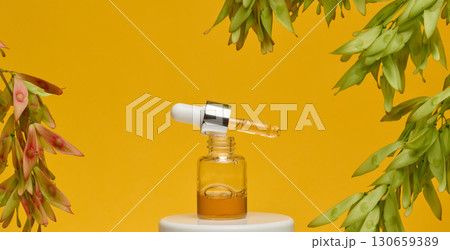 A clear glass dropper bottle with a silver cap and a white dropper dripping amber liquid, standing on a white stand. A clear glass dropper bottle with a silver cap and a white dropper dripping amber liquid, standing on a white stand. 130659389