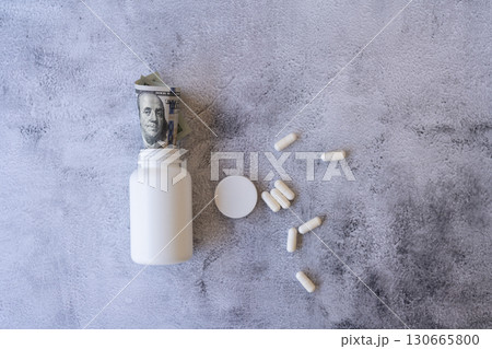 White pill bottle filled with dollar bills and scattered capsules on a gray background, concept of expensive medicine and healthcare costs. White pill bottle filled with dollar bills and scattered capsules on a gray background, concept of expensive medicine and healthcare costs. 130665800
