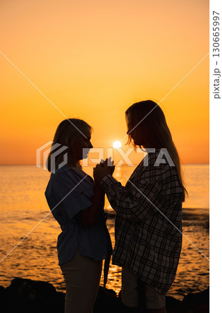 Mother and teenage daughter holding hands at sunrise on the seashore, silhouette with warm morning glow 130665997