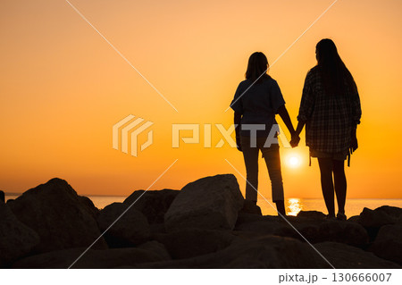 Silhouette of a middle-aged woman and a young woman holding hands on rocks by the sea at sunrise, symbolizing family love, emotional support, and the harmony of a new day Silhouette of a middle-aged woman and a young woman holding hands on rocks by the sea at sunrise, symbolizing family love, emotional support, and the harmony of a new day 130666007
