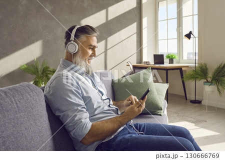 Bearded man in headphones listening to music, sitting on the sofa having rest at home. 130666769