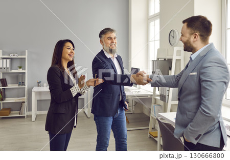 Two business partners handshaking in office, colleague applauding them Two business partners handshaking in office, colleague applauding them 130666800
