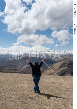 Person standing on a hill with raised arms facing Mount Elbrus on a partly cloudy day 130667521