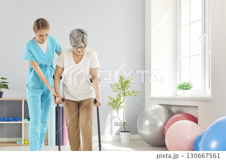 Young female caregiver helping elderly woman to walk with crutches during rehabilitation in clinic. 130667561