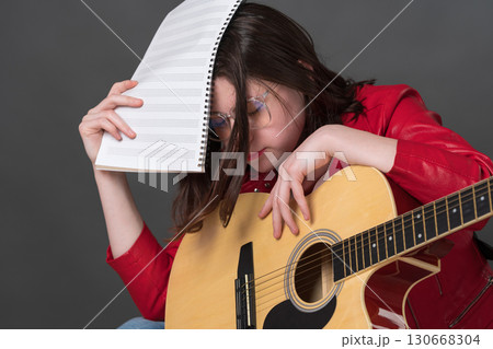 Tired and exhausted female musician with guitar and notes in hand covers face with music sheets due to fatigue from long and tiring rehearsal and learning to play classical six string acoustic guitar Tired and exhausted female musician with guitar and notes in hand covers face with music sheets due to fatigue from long and tiring rehearsal and learning to play classical six string acoustic guitar 130668304