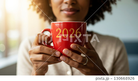 A woman holds a red mug with 2026 on it, celebrating the New Year with a warm drink and a bright, sunlit background. A woman holds a red mug with 2026 on it, celebrating the New Year with a warm drink and a bright, sunlit background. 130668926