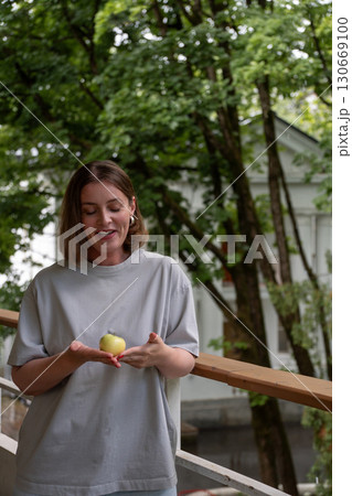 Smiling woman stands on a veranda in Peredelkino, looking down at a green apple gently held in her cupped hands Smiling woman stands on a veranda in Peredelkino, looking down at a green apple gently held in her cupped hands 130669100