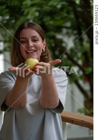 Woman gently presenting a green apple in her cupped hands, smiling with greenery in the background Woman gently presenting a green apple in her cupped hands, smiling with greenery in the background 130669123