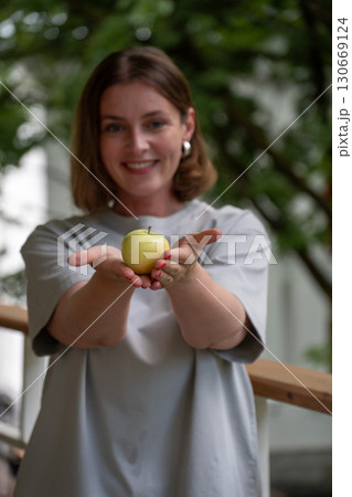 Woman gently presenting a green apple in her cupped hands, smiling with greenery in the background Woman gently presenting a green apple in her cupped hands, smiling with greenery in the background 130669124