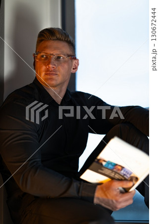 A man wearing a black shirt and glasses sits on a windowsill focused on reading a book. Soft afternoon light brightens the cozy indoor space enhancing a peaceful atmosphere. 130672444