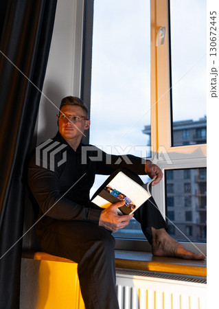 man in a black shirt and glasses sits barefoot on a windowsill deeply engrossed in a book. The backdrop shows a modern cityscape at dusk creating a serene atmosphere. 130672445