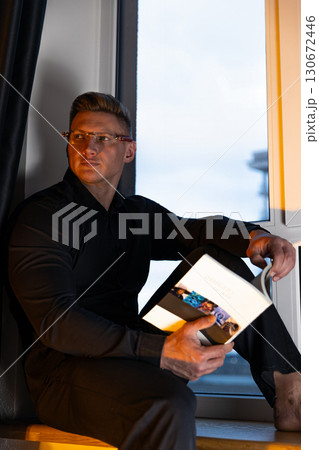 man dressed in a black shirt and glasses sits on a windowsill absorbed in reading a book. The warm glow of evening light adds a cozy atmosphere to his quiet moment. man dressed in a black shirt and glasses sits on a windowsill absorbed in reading a book. The warm glow of evening light adds a cozy atmosphere to his quiet moment. 130672446
