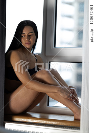 girl with long dark hair sits on a windowsill gazing thoughtfully out. The natural light illuminates her features creating a serene and reflective atmosphere. girl with long dark hair sits on a windowsill gazing thoughtfully out. The natural light illuminates her features creating a serene and reflective atmosphere. 130672461