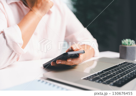 Close-up shot, woman sits at a wooden table using her smartphone, with her finger touching the phone screen as she navigates mobile banking, online shopping, and social media networks. 130676455