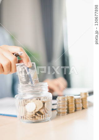 Hand places coin into saving jar, representing how smart finance, investment, banking practices turn money, cash, currency into financial wealth drives business growth and strengthens the economy. Hand places coin into saving jar, representing how smart finance, investment, banking practices turn money, cash, currency into financial wealth drives business growth and strengthens the economy. 130677898