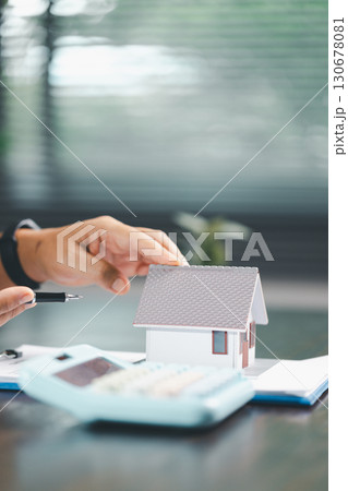 Real estate agent and customer are discussing contract to buy house, with house model in background symbolizing real estate, insurance, and loan arrangements involved in process. 130678081