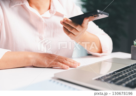 Close-up shot, woman sits at a wooden table using her smartphone, with her finger touching the phone screen as she navigates mobile banking, online shopping, and social media networks. 130678196