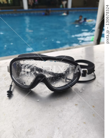 A pair of wet swimming goggles sits on a poolside table with water droplets visible on the lenses. In the blurry background, people are swimming and enjoying the pool. 130678324