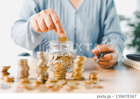 Hand places coin into saving jar, representing how smart finance, investment, banking practices turn money, cash, currency into financial wealth drives business growth and strengthens the economy. Hand places coin into saving jar, representing how smart finance, investment, banking practices turn money, cash, currency into financial wealth drives business growth and strengthens the economy. 130678367