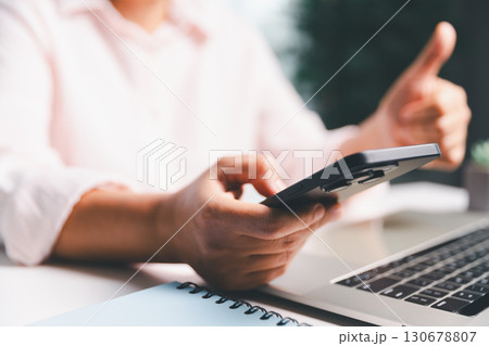 Close-up shot, woman sits at a wooden table using her smartphone, with her finger touching the phone screen as she navigates mobile banking, online shopping, and social media networks. 130678807